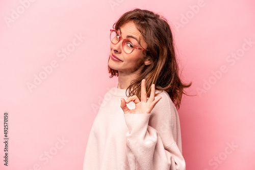 Young caucasian woman isolated on pink background winks an eye and holds an okay gesture with hand. photo