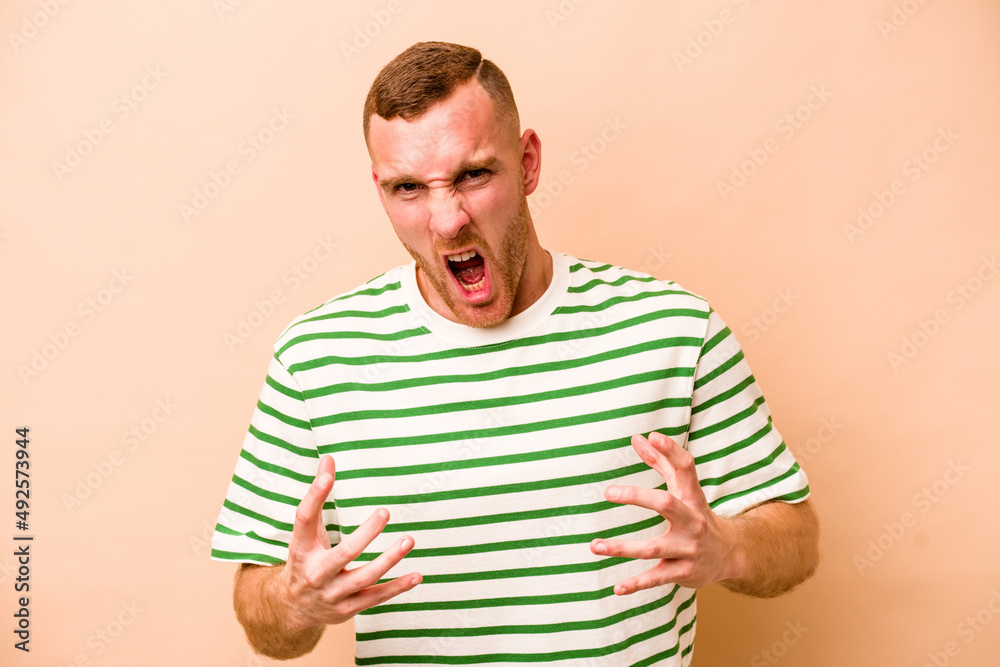 Young caucasian man isolated on beige background upset screaming with tense hands. Young caucasian man isolated on beige background upset screaming with tense hands.