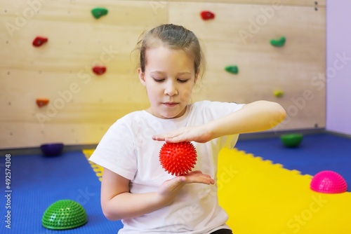 a little girl in white holds a red ball with spikes and massages her hands in a children's center so as not to get sick