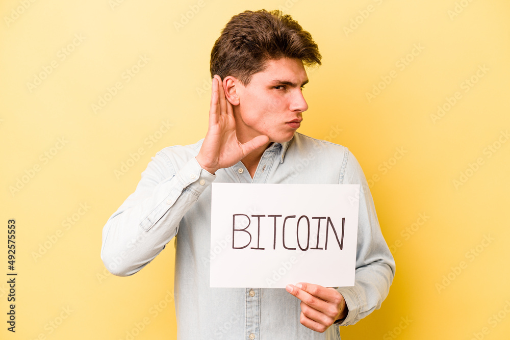 Young caucasian man holding bitcoin placard isolated on yellow background trying to listening a gossip. Young caucasian man holding bitcoin placard isolated on yellow background trying to listening a gossip.
