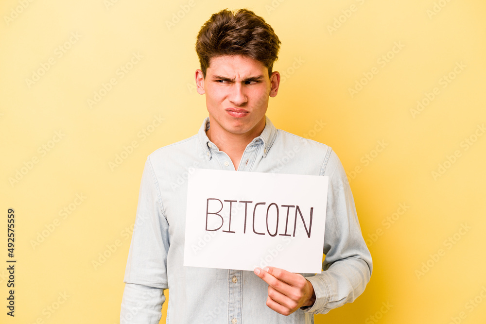 Young caucasian man holding bitcoin placard isolated on yellow background confused, feels doubtful and unsure. Young caucasian man holding bitcoin placard isolated on yellow background confused, feels doubtful and unsure.