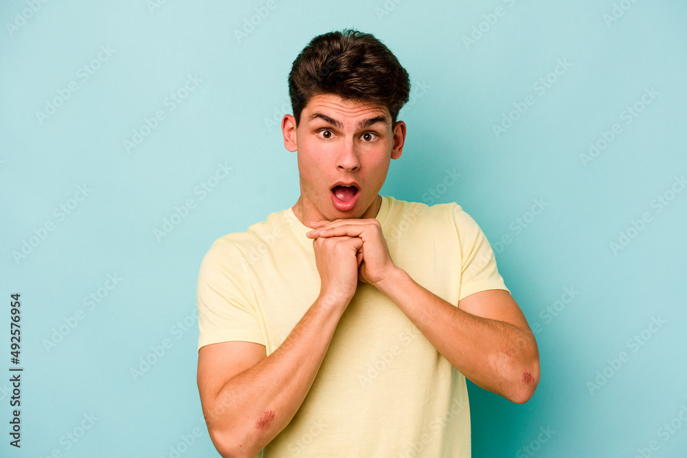 Young caucasian man isolated on blue background praying for luck, amazed and opening mouth looking to front.