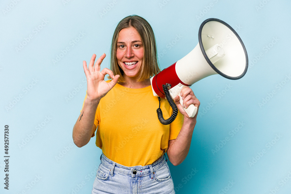 Naklejka premium Young caucasian woman holding a megaphone isolated on blue background cheerful and confident showing ok gesture.