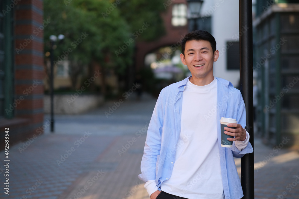 smiling Asian young man holding a cup of coffee on the street, looking at camera smiling Asian young man holding a cup of coffee on the street, looking at camera