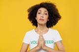 Young woman of African American ethnicity wears white volunteer t-shirt hold hand folded in prayer gesture, beg isolated on plain yellow background. Voluntary free work assistance help grace concept.