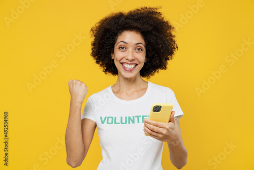 Young woman of African American ethnicity in white volunteer t-shirt hold use mobile cell phone do winner gesture isolated on plain yellow background Voluntary free work assistance help grace concept.