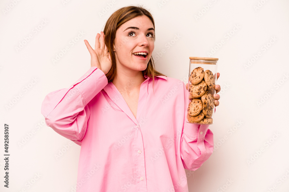 Young caucasian woman holding cookies jar isolated on white background trying to listening a gossip. Young caucasian woman holding cookies jar isolated on white background trying to listening a gossip.