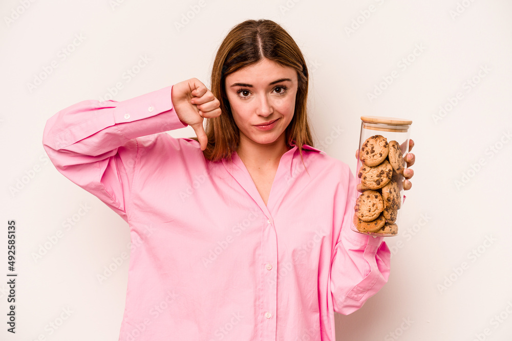 Young caucasian woman holding cookies jar isolated on white background showing a dislike gesture, thumbs down. Disagreement concept. Young caucasian woman holding cookies jar isolated on white background showing a dislike gesture, thumbs down. Disagreement concept.
