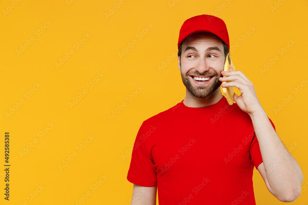 Professional delivery guy employee man in red cap T-shirt uniform workwear work as dealer courier speak talk on mobile cell phone isolated on plain yellow background studio portrait Service concept.