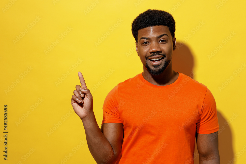 Young smiling man of African American ethnicity 20s wear orange t-shirt point index finger aside on workspace area mock up isolated on plain yellow background studio portrait People lifestyle concept Young smiling man of African American ethnicity 20s wear orange t-shirt point index finger aside on workspace area mock up isolated on plain yellow background studio portrait People lifestyle concept