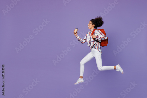 Full size young girl woman of African American ethnicity teen student in shirt hold backpack jump high hurry up run fast drink coffee isolated on plain purple background. Education in college concept.