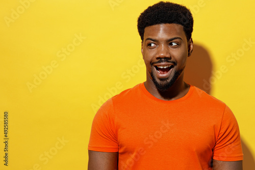 Young surprised man of African American ethnicity 20s wear orange t-shirt look aside on workspace area mock up copy space isolated on plain yellow background studio portrait. People lifestyle concept.