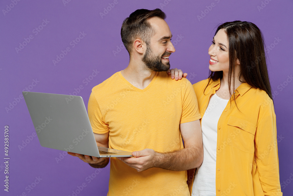 Young smiling happy couple two friends family man woman together in yellow casual clothes hold use work on laptop pc computer look to each other isolated on plain violet background studio portrait. Young smiling happy couple two friends family man woman together in yellow casual clothes hold use work on laptop pc computer look to each other isolated on plain violet background studio portrait.