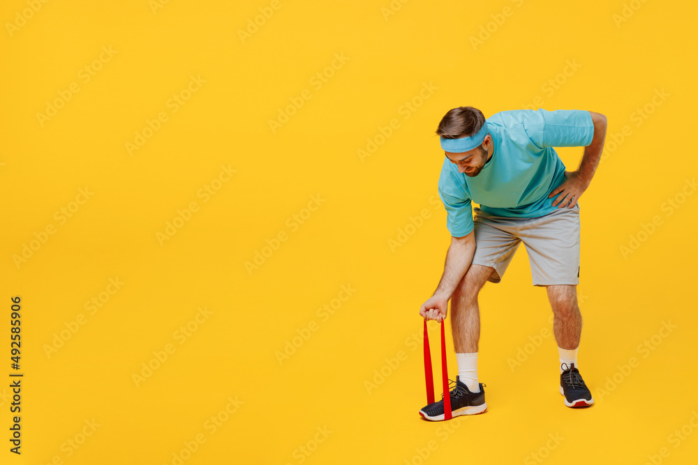 Fototapeta premium Full body side view young fitness trainer instructor sporty man sportsman wear headband blue t-shirt use fitness elastic bands for hands isolated on plain yellow background. Workout sport concept