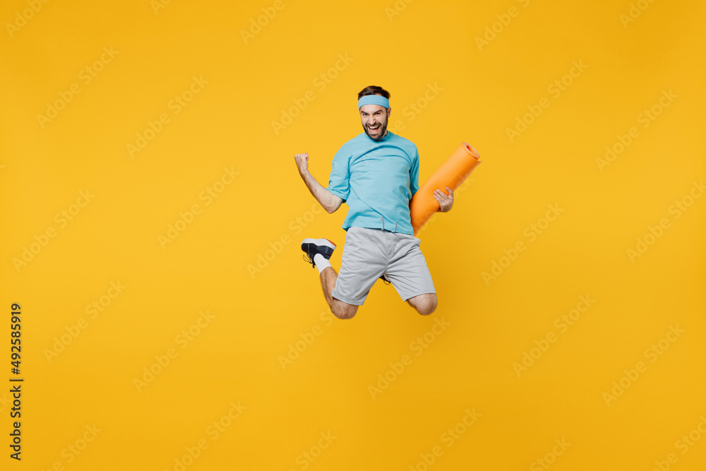 Full body overjoyed young fitness trainer instructor sporty man sportsman in headband blue t-shirt hold yoga mat jump high do winner gesture isolated on plain yellow background. Workout sport concept