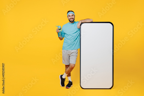 Full body young fitness trainer instructor sporty man sportsman in headband blue t-shirt big screen mobile cell phone with copy space mockup isolated on plain yellow background. Workout sport concept.