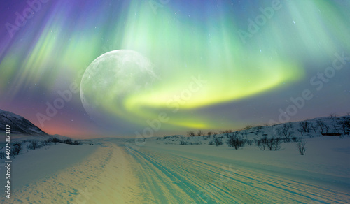 Beautiful winter landscape with snow and ice covered road full moon in the background - Northern lights (Aurora borealis) in the sky over Tromso, Norway