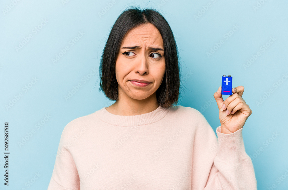 Young hispanic woman holding batteries isolated on blue background confused, feels doubtful and unsure. Young hispanic woman holding batteries isolated on blue background confused, feels doubtful and unsure.