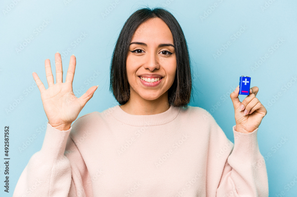 Young hispanic woman holding batteries isolated on blue background smiling cheerful showing number five with fingers. Young hispanic woman holding batteries isolated on blue background smiling cheerful showing number five with fingers.