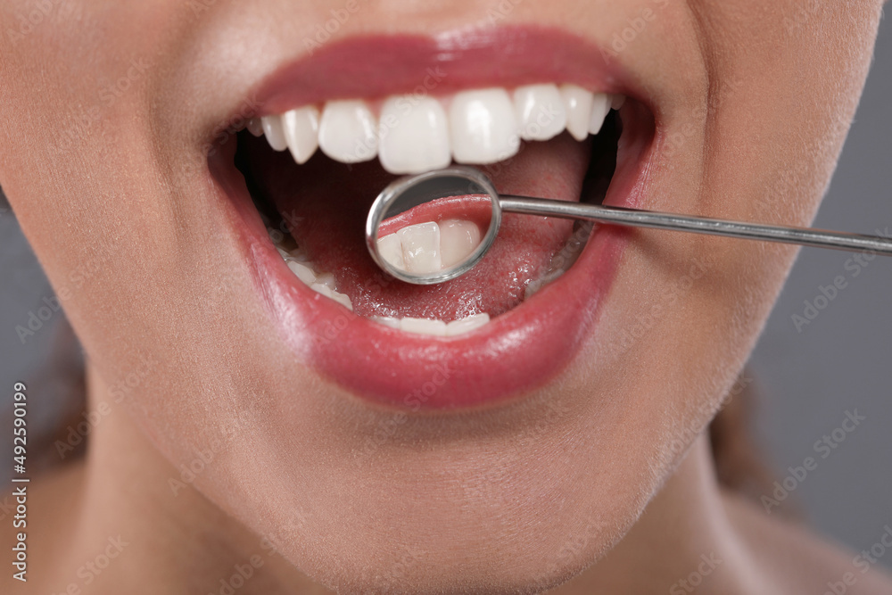 Obraz premium Examining woman's teeth with dentist's mirror on grey background, closeup