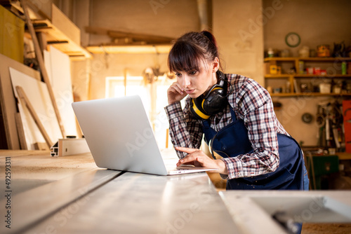 Craftswoman using laptop with blueprints in her carpentry workshop