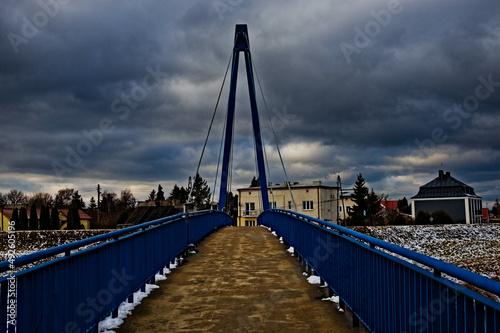 Wiszący most " huśtany " - poręcze przęsło , pylon i liny nośne - pomalowany na niebiesko na tle mocno zachmurzonego nieba .