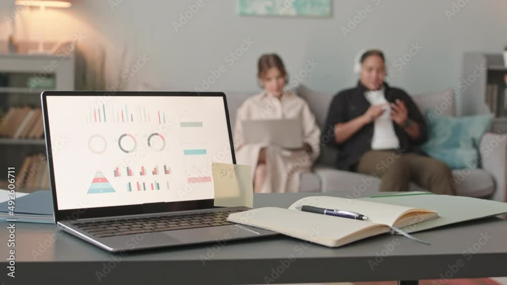 Slowmo of business diagrams on laptop screen at foreground and interracial lesbian couple sitting on sofa at background. One of women working on laptop while her partner listening to music nearby Slowmo of business diagrams on laptop screen at foreground and interracial lesbian couple sitting on sofa at background. One of women working on laptop while her partner listening to music nearby