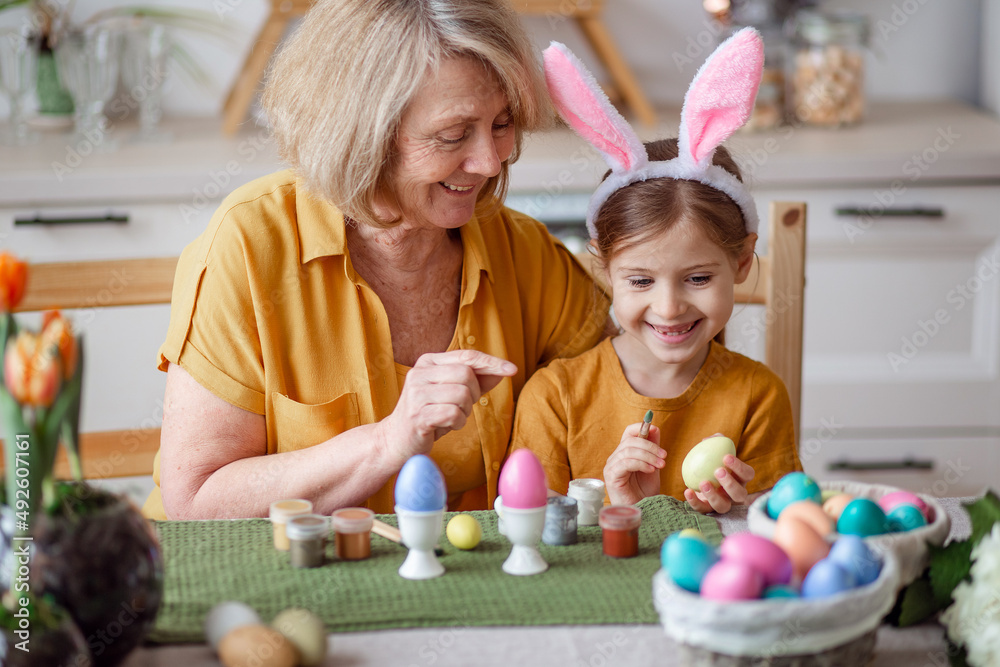 Obraz premium Happy easter family elderly grandmother and little granddaughter with rabbit ears are preparing for the holiday to paint eggs