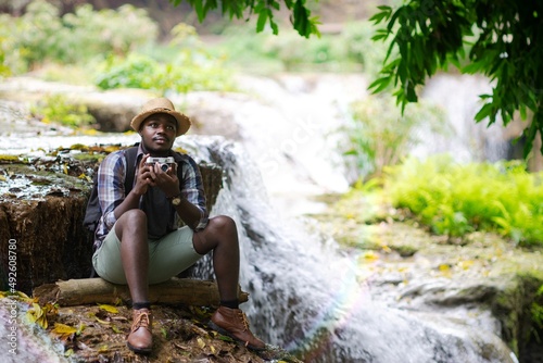 Fototapeta Naklejka Na Ścianę i Meble -  African Man Traveller holding a film camera with backpack sitting and relaxing freedom at the waterfall.