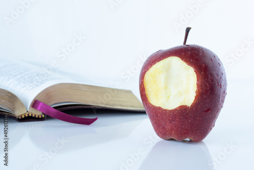 Red bitten apple with an open Holy Bible Book on white background. The Christian biblical concept of the biblical story in Genesis of forbidden fruit, temptation, and disobedience to God. A closeup. photo
