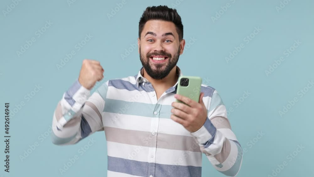 Happy young bearded brunet man 20s wears striped shirt hold use mobile cell phone say yes found out great big win news do winner gesture isolated on plain pastel light blue background studio portrait Happy young bearded brunet man 20s wears striped shirt hold use mobile cell phone say yes found out great big win news do winner gesture isolated on plain pastel light blue background studio portrait