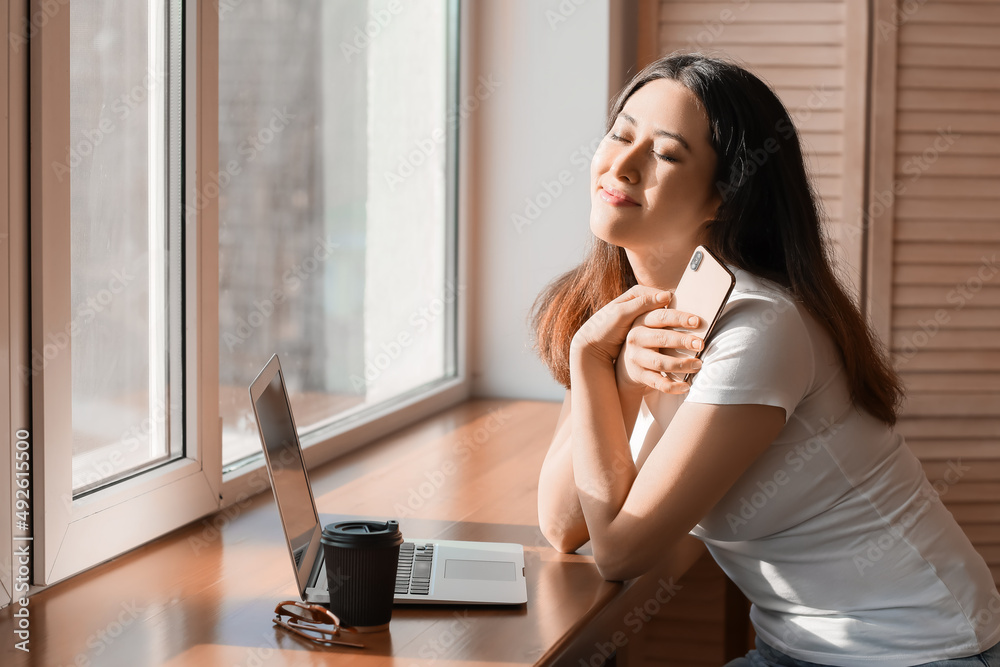 Happy young Asian woman with mobile phone and laptop near window