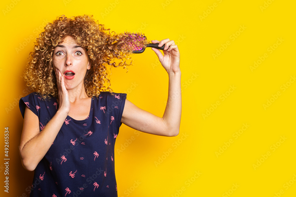 surprised young woman combing tangled curly hair on yellow background surprised young woman combing tangled curly hair on yellow background