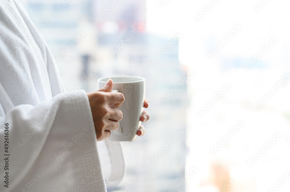 Side view of woman in bathrobe standing near the window while holding white cup of coffee and enjoy wonderful morning in the hotel, detaching from work. Side view of woman in bathrobe standing near the window while holding white cup of coffee and enjoy wonderful morning in the hotel, detaching from work.
