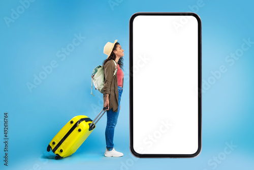 Black female traveler with suitcase and backpack looking at big smartphone with mockup for online travel agency website