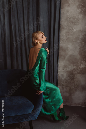 A beautiful blonde woman in an elegant green silk dress with an open back sits on a blue armchair against a dark background. Beauty industry. Soft selective focus.