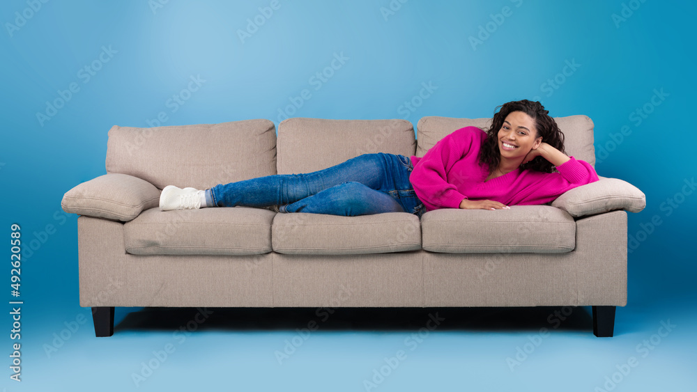 Beautiful happy young black woman in casual wear lying on sofa and smiling at camera on blue studio background Beautiful happy young black woman in casual wear lying on sofa and smiling at camera on blue studio background