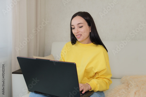 The brunette blogger smiles and looks at the laptop monitor while sitting on the couch at home. © Anna