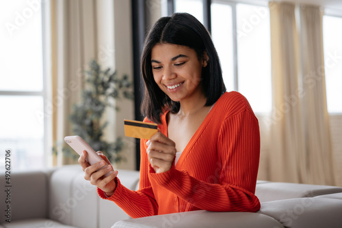 Smiling African American woman  holding credit card using mobile phone shopping online, check card balance. Mobile banking. Beautiful female ordering food online looking at screen sitting at home 