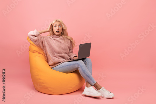 Deadline concept. Shocked young woman sitting in beanbag chair with laptop and grabbing her head, pink background