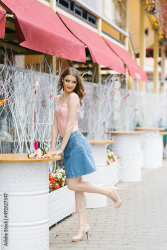 Cute young woman in a denim skirt and pink top stands near a metal white barrel on a walk in the city © Sunshine