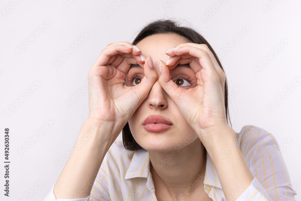 Close up portrait of attractive quirky young woman making binoculars with hands showing ok gesture on white studio background.