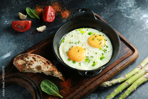 Breakfast. Fried eggs in a cast-iron pan with fried bread, beans, asparagus, tomatoes, herbs on a wooden board on a dark table. Rustic. Background image, copy space.