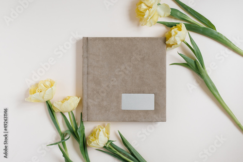 Beige fabric family or wedding photo album with empty space for text surrounded by yellow tulips on a light background. Family photo archive