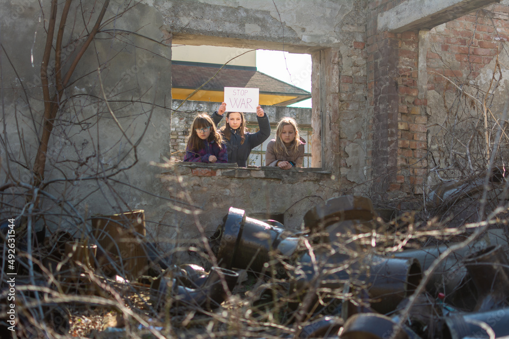 Three desperate girls in winter jackets standing on ruins of destroyed buildings in war zone, with Board No war, Stop war. Refugee children. The war in the Ukraine, combat actions.