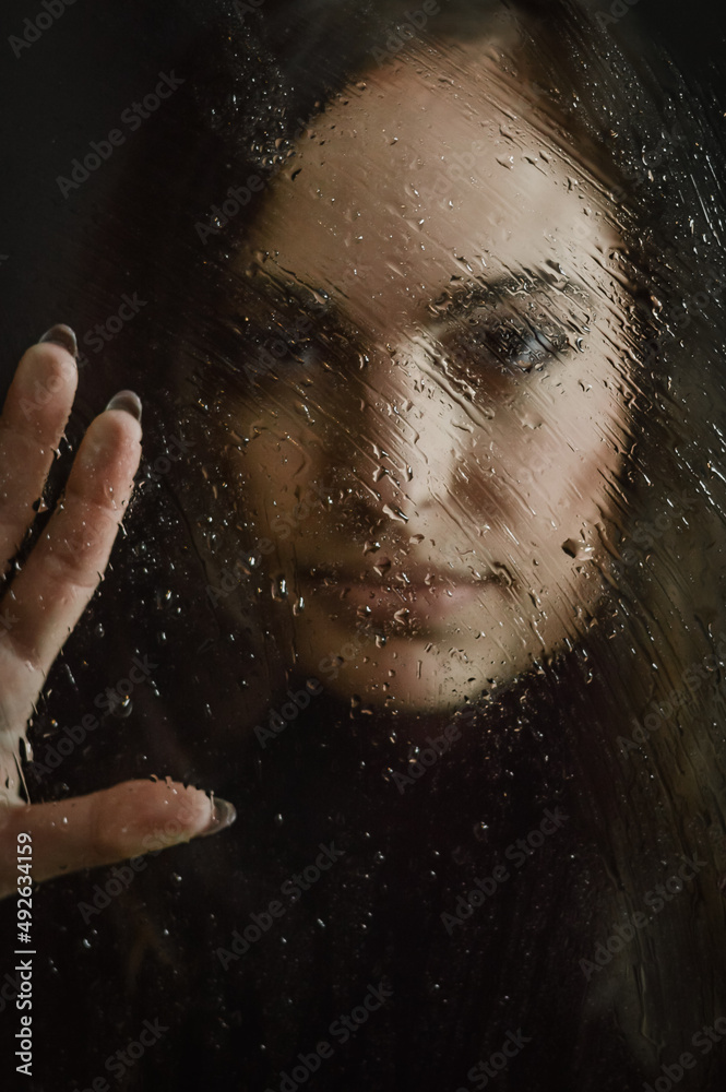portrait of a beautiful girl with long hair leaning a hand against a glass covered with water droplets, focus on the glass and the droplets