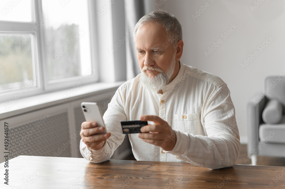 Man pays for an online purchase on phone with a bank card. An old gray man with a beard at the table does online shopping. Close up.