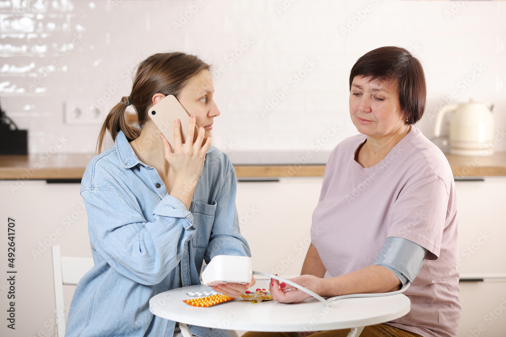 worried young woman calling doctor for her senior mother at home. sick senior woman with medication sitting at the table and measuring blood pressure. medicine and health concept worried young woman calling doctor for her senior mother at home. sick senior woman with medication sitting at the table and measuring blood pressure. medicine and health concept