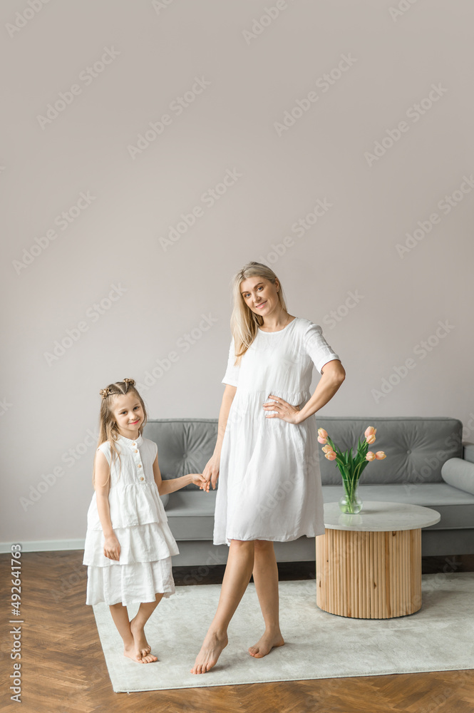 Mom and daughter hug at home near the couch. A young woman and a girl in white linen dresses. Mom and daughter hug at home near the couch. A young woman and a girl in white linen dresses.