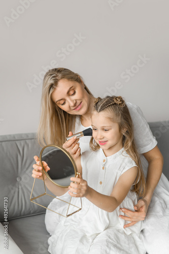 Mom and daughter play with cosmetics in a cozy home. A young woman and a girl in white linen dresses sit on the couch and make up for each other.
